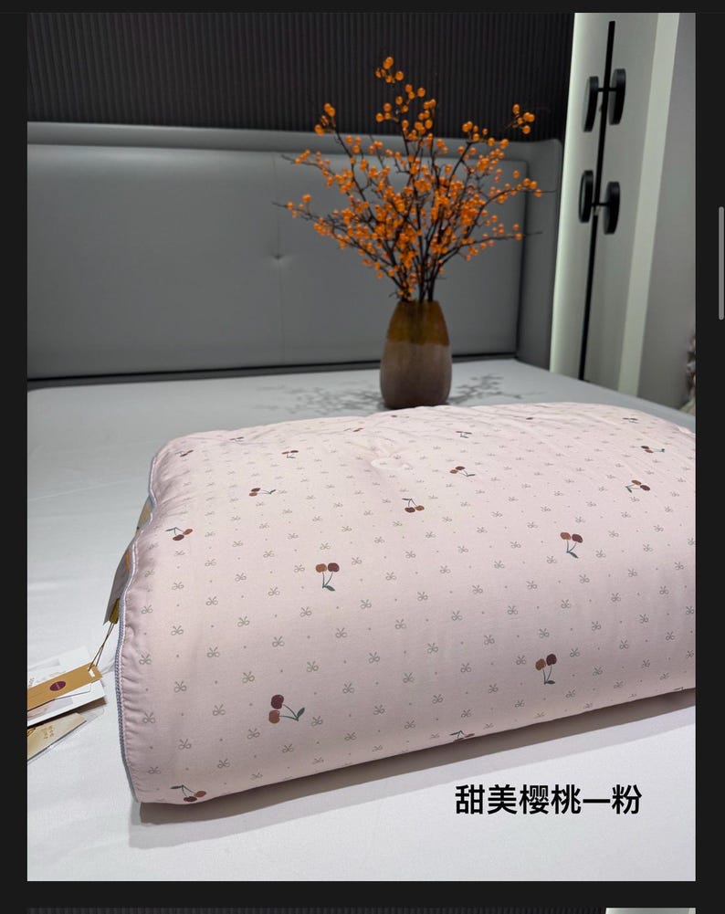 May include: A light pink pillow with a cherry pattern. The pillow is on a bed with a gray headboard. A vase with orange branches sits on the bed. The text on the pillow reads "Sweet Cherry Pink".