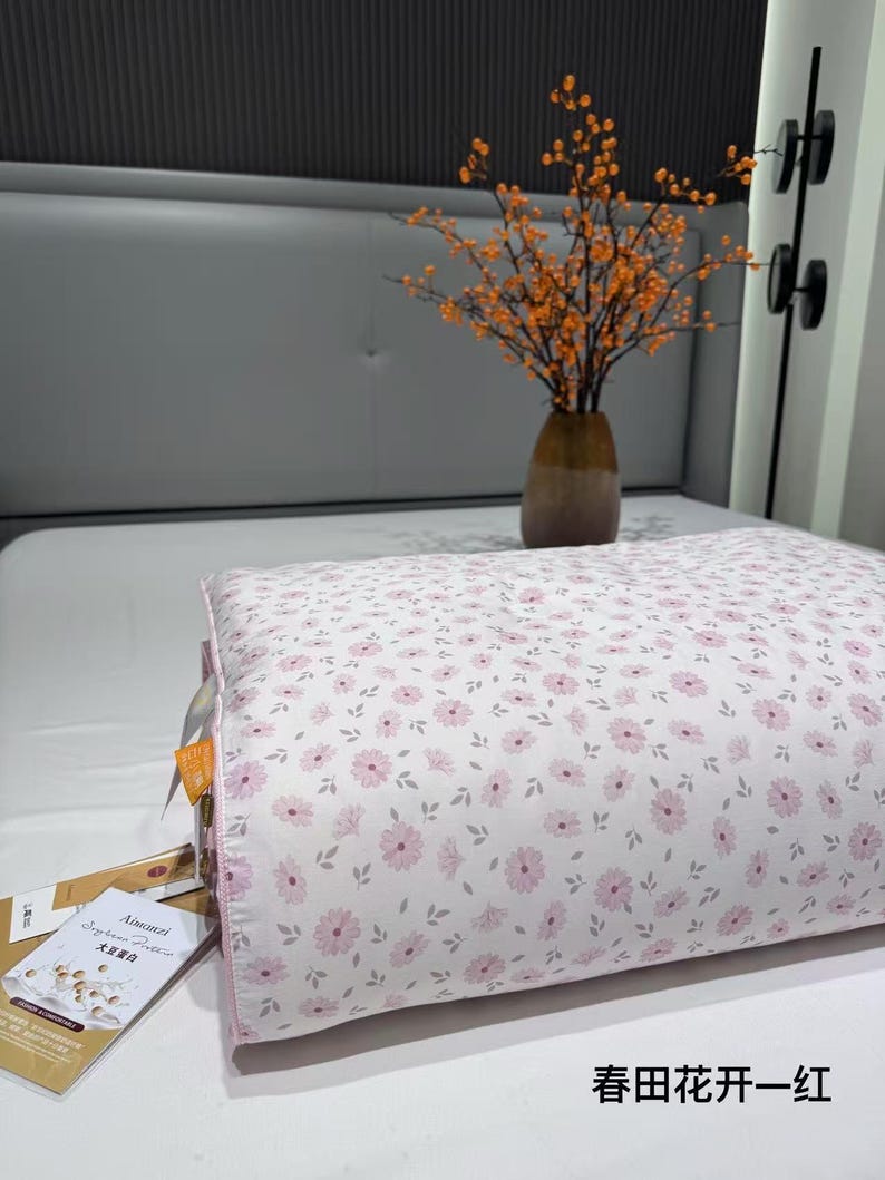 May include: A floral-patterned pillow with pink flowers and gray leaves on a white background. The pillow has a pink border and is on a white bed. The text "春田花开一红" is visible.