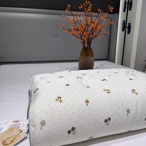 May include: A white pillow with a pattern of brown and tan cherries and trees. The pillow is on a bed with a gray headboard. A vase of orange flowers sits on the bed. The text "cherry cloth Ding-green" is at the bottom.