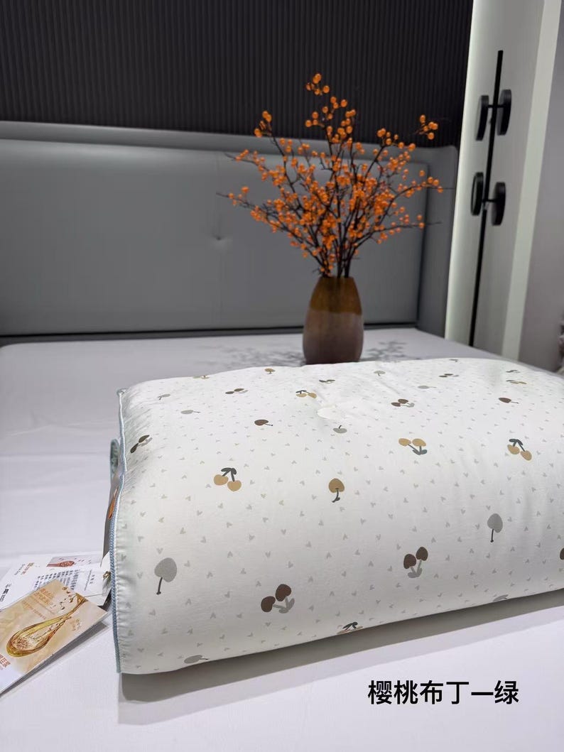 May include: A white pillow with a pattern of brown and tan cherries and trees. The pillow is on a bed with a gray headboard. A vase of orange flowers sits on the bed. The text "cherry cloth Ding-green" is at the bottom.