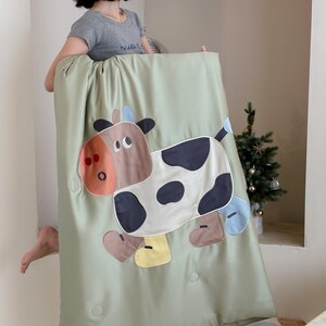 May include: A light green blanket featuring a large, cartoon cow applique. The cow has brown, black, and white patches, with accents of orange, blue, and yellow. The blanket has a quilted appearance with several button details.