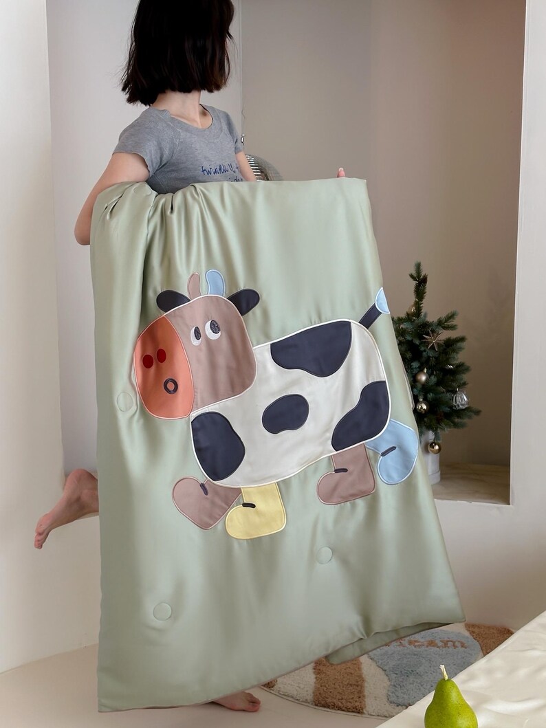 May include: A light green blanket featuring a large, cartoon cow applique. The cow has brown, black, and white patches, with accents of orange, blue, and yellow. The blanket has a quilted appearance with several button details.