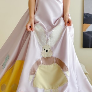 May include: A light purple dress with a large bunny applique and mushroom details. The bunny has a cream-colored face and ears, a brown body, and a yellow dress. The dress has the text "Gonna Kill You" on the top.
