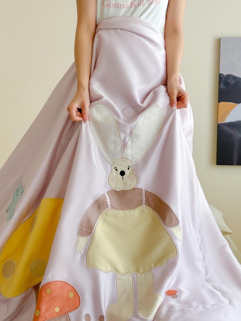 May include: A light purple dress with a large bunny applique and mushroom details. The bunny has a cream-colored face and ears, a brown body, and a yellow dress. The dress has the text "Gonna Kill You" on the top.