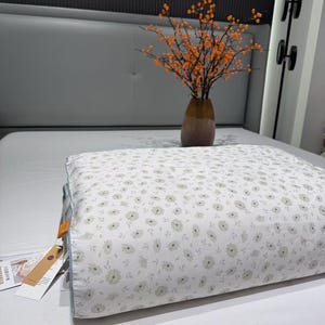 May include: A white pillow with a floral pattern of small, light green flowers. The pillow is on a white bed, with a vase of orange-berried branches in the background. The text "Spring Field Flowers - Green" is visible.