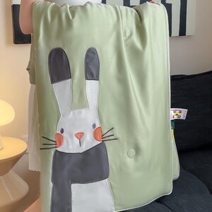 May include: A light green blanket featuring a large, embroidered cartoon rabbit with a white face, gray scarf, and orange cheeks. The blanket has a white border and a button closure. The text on the tank top says "My Daddy's Gonna Kill You".
