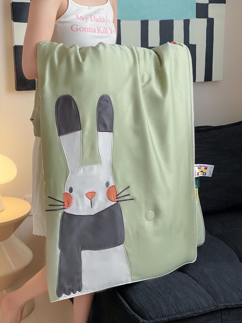 May include: A light green blanket featuring a large, embroidered cartoon rabbit with a white face, gray scarf, and orange cheeks. The blanket has a white border and a button closure. The text on the tank top says "My Daddy's Gonna Kill You".