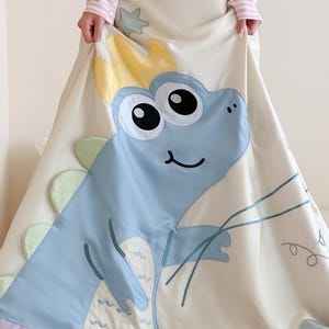 May include: A cream-colored, satin-like blanket featuring a large, light blue cartoon dinosaur with a yellow crown. The dinosaur has large black eyes and a smiling mouth. The blanket has a lavender border and green and yellow accents.