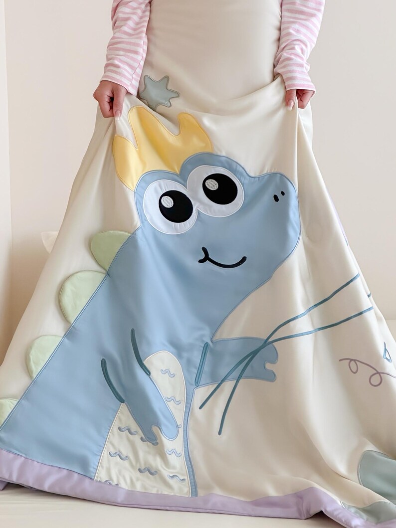May include: A cream-colored, satin-like blanket featuring a large, light blue cartoon dinosaur with a yellow crown. The dinosaur has large black eyes and a smiling mouth. The blanket has a lavender border and green and yellow accents.