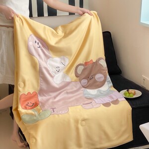 May include: Yellow blanket with cartoon dinosaur, bear, and flower appliqu&eacute;s. The dinosaur is pink, the bear is brown, and the flower is orange. The blanket has a button closure and is being held up by a person.
