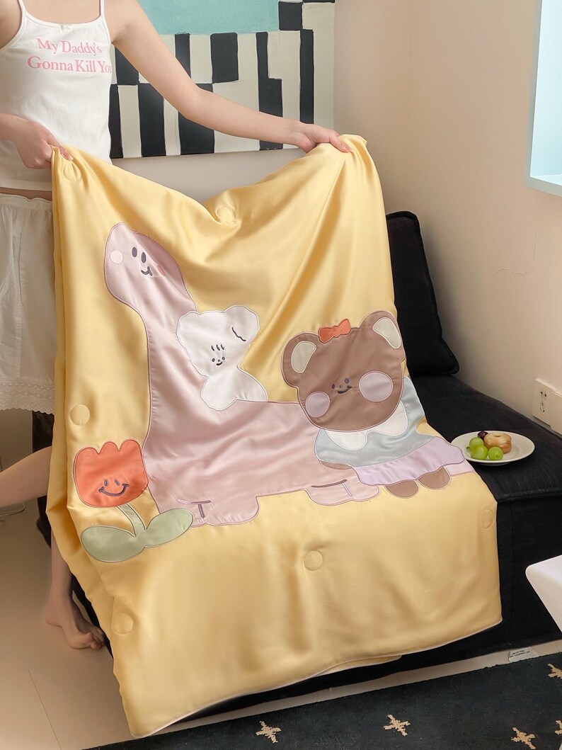 May include: Yellow blanket with cartoon dinosaur, bear, and flower appliqu&eacute;s. The dinosaur is pink, the bear is brown, and the flower is orange. The blanket has a button closure and is being held up by a person.