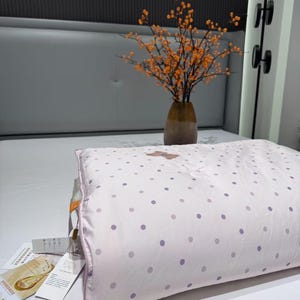 May include: A light pink pillow with a polka dot pattern in shades of purple. The pillow has a light pink border and a tag attached. A vase with orange flowers sits in the background.