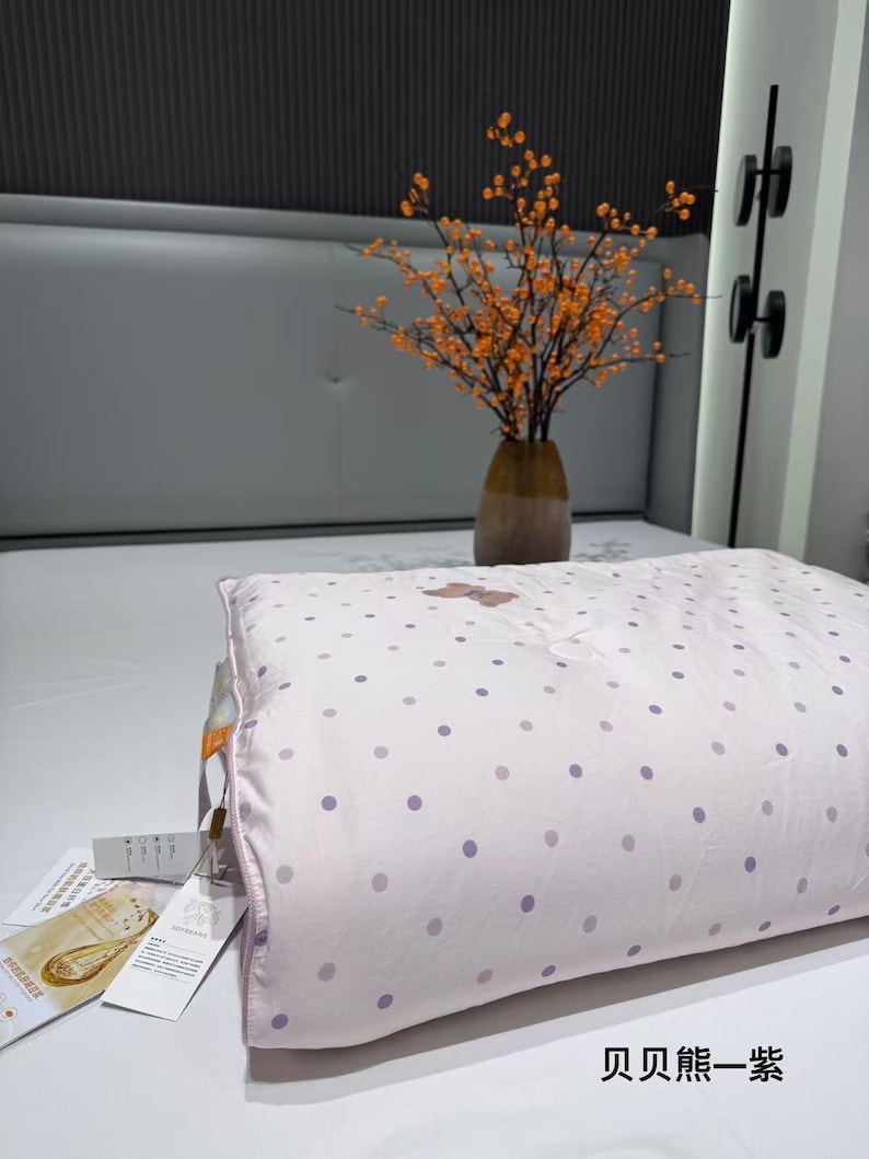 May include: A light pink pillow with a polka dot pattern in shades of purple. The pillow has a light pink border and a tag attached. A vase with orange flowers sits in the background.