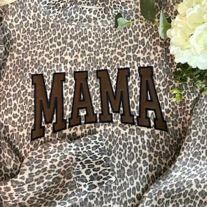 Peut inclure: Un sweat-shirt à manches longues avec un motif léopard gris et noir sur l'ensemble. Le vêtement présente un col rond et des poignets côtelés. Le mot MAMA est inscrit au centre en grandes lettres marron foncé avec des contours noirs.