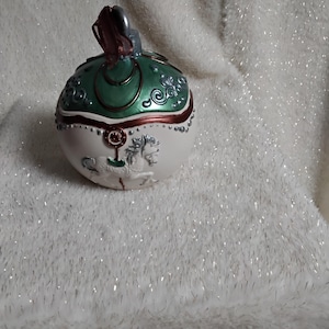 Op de afbeelding: Een decoratieve, eivormige sieradendoos met een witte basis en een groene bovenkant. De bovenkant heeft sierlijke zilveren details en een rode accent. De basis heeft een paardenreliëf. Op een witte, pluizige achtergrond.