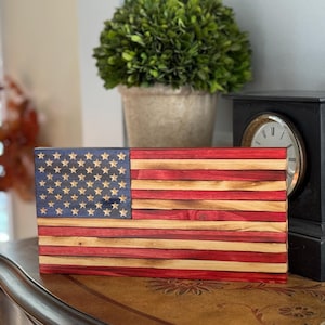 May include: Wooden American flag with a blue star field and red and natural wood stripes. The flag is rectangular and appears to be handmade. The stars are light-colored and the stripes alternate between red and natural wood.