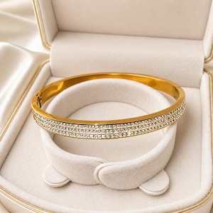 May include: A gold-toned hinged bangle bracelet featuring three parallel rows of clear, sparkling crystals embedded along the outer surface. The bracelet has a smooth, polished metal finish and a secure clasp mechanism visible on the side.