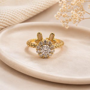 May include: A gold-toned ring featuring a central round clear stone surrounded by a halo of smaller clear stones. The band has a textured cross-hatch pattern and is topped with two small, upright, cream-colored rabbit ears behind the stone setting.