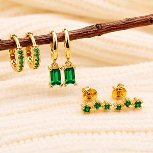 May include: A set of gold-toned jewelry featuring two small hoop earrings with green stones, two hoop earrings with dangling rectangular green stones, and two stud earrings each with three small green stones arranged in a horizontal row.