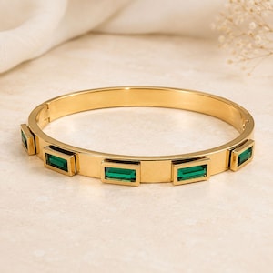 May include: A gold-toned hinged bangle bracelet featuring four rectangular, emerald-green baguette-cut stones set at intervals along the outer surface of the polished metal band, which includes a secure clasp mechanism on the side.