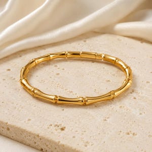 Op de afbeelding: Een gepolijste goudkleurige armband met een herhalend, op bamboe geïnspireerd ontwerp met gesegmenteerde knopen. Het cirkelvormige sieraad heeft een gladde, reflecterende metalen afwerking en een doorlopende, uniforme structuur over de gehele band.