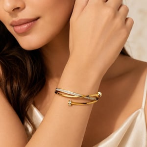 Könnte beinhalten: Ein goldfarbenes Bypass-Armband mit einem polierten Metallband, das sich um das Handgelenk legt. Ein Ende des Bandes ist glatt und abgerundet, während das andere Ende mit einer Reihe kleiner, klarer, funkelnder Steine auf der Oberfläche verziert ist.