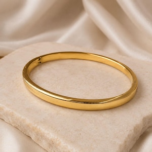 May include: A polished gold-tone metal bangle bracelet with a smooth, rounded surface and a visible hinged clasp mechanism. The bracelet features a continuous, solid band design with a reflective finish, showcasing a clean and simple circular shape.