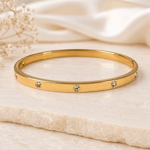 Puede incluir: Una pulsera rígida de metal en tono dorado con un acabado liso y pulido, que presenta una serie de cristales transparentes de corte redondo espaciados uniformemente a lo largo de la superficie exterior y un cierre de bisagra lateral.