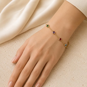 May include: A gold chain bracelet featuring four evenly spaced, round, bezel-set gemstones in green, purple, red, and blue. The delicate metal links connect each circular stone setting, creating a simple, colorful, and refined piece of jewelry.