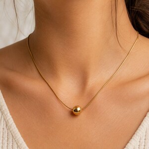 May include: A gold-toned necklace featuring a thin, smooth snake chain and a single, polished spherical bead pendant centered on the chain. The necklace rests against the neck, showcasing a reflective, metallic finish on both the bead and chain.