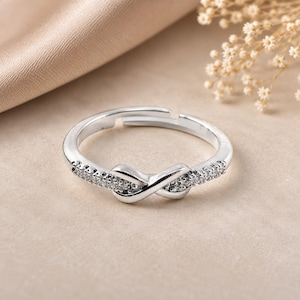 Peut inclure: Une bague argentée ajustable ornée d'un symbole infini en son centre. L'anneau est agrémenté de petites pierres claires et scintillantes incrustées sur les côtés, menant au motif central, offrant un aspect poli et très réfléchissant.