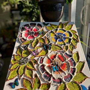 May include: A textured floral artwork on a white canvas. The piece features three large, colourful flowers with intricate details in red, blue, yellow, and white. Green leaves and butterfly accents surround the flowers, creating a vibrant, dimensional effect.