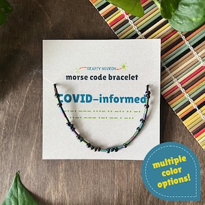 May include: A Morse code bracelet featuring iridescent, multi-colored beads on a black cord, displayed on a white card labeled Crafty Neuron, morse code bracelet, COVID-informed, and a green Morse code pattern, with a blue sticker stating multiple color options.