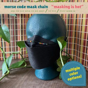 May include: A black face mask worn on a dark green mannequin head, featuring a beaded chain with Morse code patterns. The text reads: morse code mask chain, masking is hot, and multiple color options. The chain has small, colorful beads.