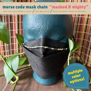 May include: A black face mask worn on a dark teal mannequin head, featuring a beaded chain with gold and light blue beads arranged in a Morse code pattern. The text "morse code mask chain "masked & mighty"" and "multiple color options!" appear.