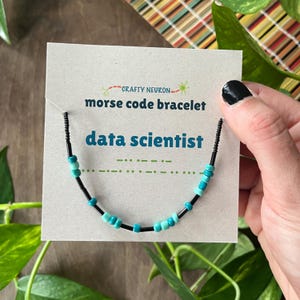 May include: A Morse code bracelet featuring alternating black and turquoise beads on a light gray display card. The card displays the text Crafty Neuron, morse code bracelet, and data scientist in blue and black, with green Morse code dots and dashes.
