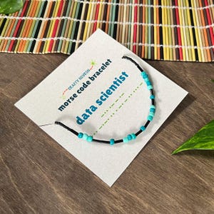 May include: A Morse code bracelet on a white display card featuring the text Crafty Neuron, morse code bracelet, and data scientist. The bracelet consists of black string with alternating small turquoise and light blue beads arranged in a pattern.