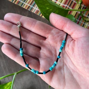 May include: A beaded bracelet featuring a repeating pattern of small black cylindrical beads interspersed with round turquoise and light blue beads. The bracelet includes a silver-toned metal lobster claw clasp at one end for secure fastening.