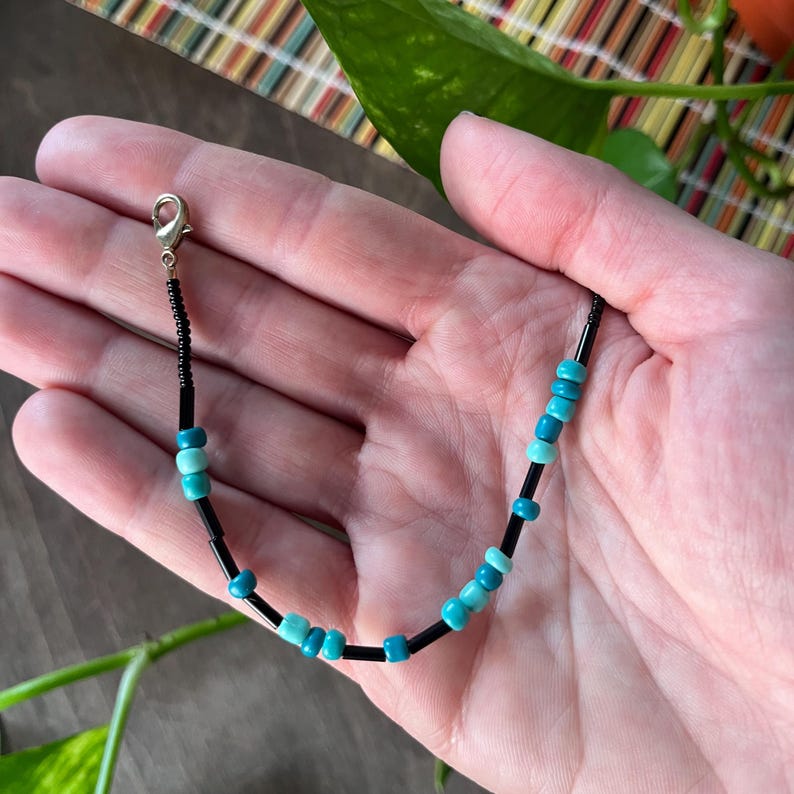 May include: A beaded bracelet featuring a repeating pattern of small black cylindrical beads interspersed with round turquoise and light blue beads. The bracelet includes a silver-toned metal lobster claw clasp at one end for secure fastening.