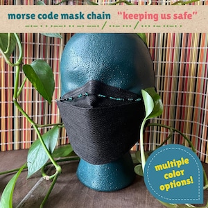 May include: A black face mask featuring a beaded chain with Morse code patterns spelling "keeping us safe" in teal and black beads. The mask is displayed on a dark teal mannequin head, with text overlays stating "morse code mask chain" and "multiple color options!"