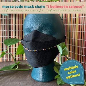 May include: A black face mask worn on a dark teal mannequin head, featuring a beaded chain with the Morse code for 'i believe in science'. The chain consists of small, multicolored beads on a thin black cord. Text reads 'morse code mask chain i believe in science' and 'multiple color options'.