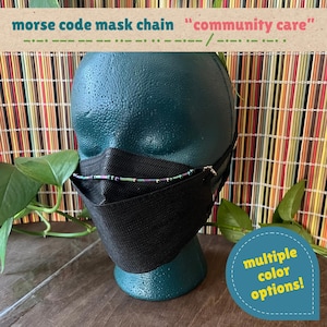 May include: A black face mask worn on a dark teal mannequin head, featuring a beaded chain with Morse code patterns. The text reads morse code mask chain community care and multiple color options. The beads are iridescent purple and green.