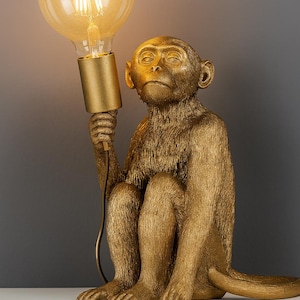 Puede incluir: Lámpara dorada con forma de mono sosteniendo una gran bombilla encendida. El mono está sentado con las piernas recogidas, y la bombilla emite un brillo cálido. La lámpara tiene un acabado texturizado y un diseño moderno.
