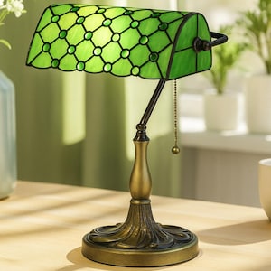 Puede incluir: Una lámpara de escritorio de vidrieras verdes con una base de bronce y una cadena de tracción. La pantalla es de color verde vibrante con un patrón geométrico. La lámpara está sobre una superficie de madera.
