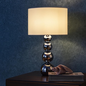 May include: A stylish table lamp featuring a dark brown, spherical base and a cream-colored rectangular lampshade. The lamp is placed on a dark wooden surface, alongside a book and a wooden sculpture.