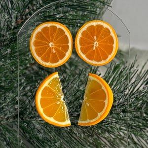 May include: A clear acrylic display stand featuring two orange slices and two orange wedges. The stand is set against a backdrop of green pine branches with a dusting of white. The orange slices are bright orange.