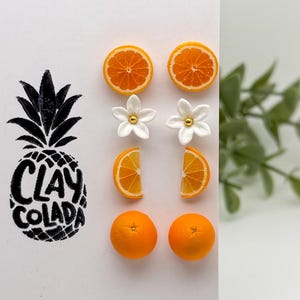May include: A white card with a black pineapple illustration and the text "CLAY COLADA." The card displays various orange-themed clay earrings, including orange slices, whole oranges, and flower designs. The earrings are arranged on the card.