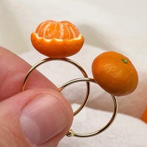 Puede incluir: Dos anillos dorados, cada uno adornado con una fruta naranja en miniatura. Un anillo presenta un segmento de naranja pelado, el otro una naranja entera. Los anillos se sostienen sobre un fondo blanco.