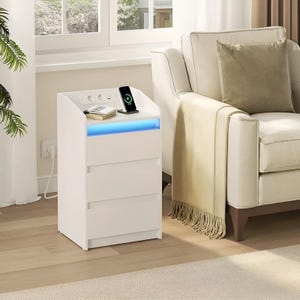 Pode incluir: Uma mesa de cabeceira branca com duas gavetas e uma superfície superior. A parte superior tem uma estação de carregamento com um telefone e um livro. Uma faixa de luz LED azul ilumina a frente. A mesa de cabeceira está ao lado de um sofá branco.