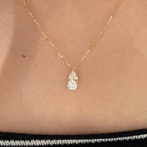 May include: A delicate gold necklace with a pear-shaped pendant set with sparkling white gemstones. The pendant is suspended from a fine gold chain. The necklace is shown against a black and white striped background.